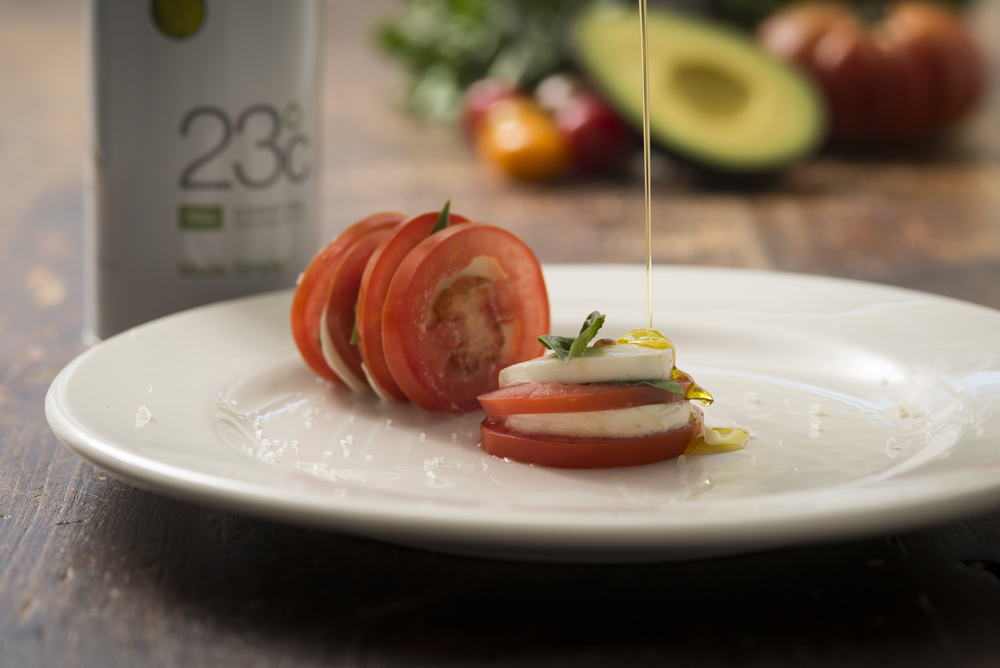 Olive oil being drizzled over tomato, mozzarella, and basil on a white plate with avocado in the background